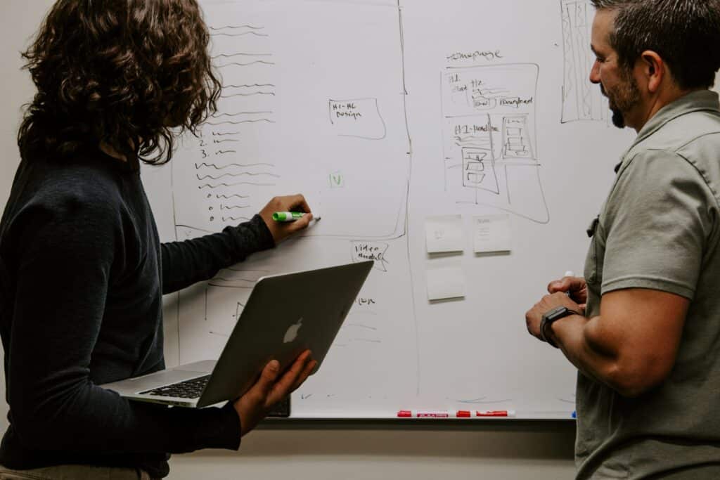 man wearing gray polo shirt beside another guy in white shirt next to a dry-erase board